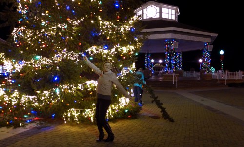 A woman poses joyfully by a brightly lit Christmas tree at night, while a person decorates nearby.