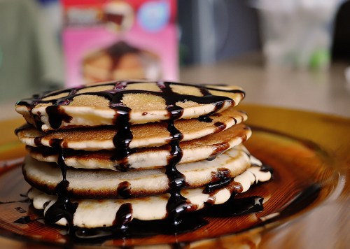 A stack of pancakes drizzled with chocolate syrup on a brown plate, with a blurred background.