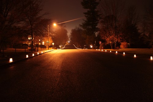 A quiet, dimly lit street at night, lined with glowing lanterns along the edges. Trees and houses are faintly visible.