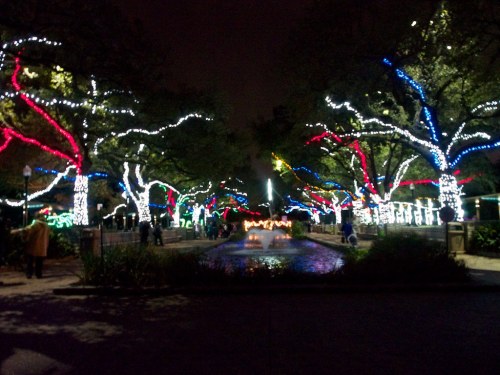 Colorful holiday lights adorn trees lining a pathway, reflecting on a calm waterway at night.