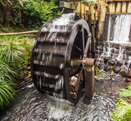 A wooden water wheel turns in a lush, green setting, with water cascading over it and rocks visible below.