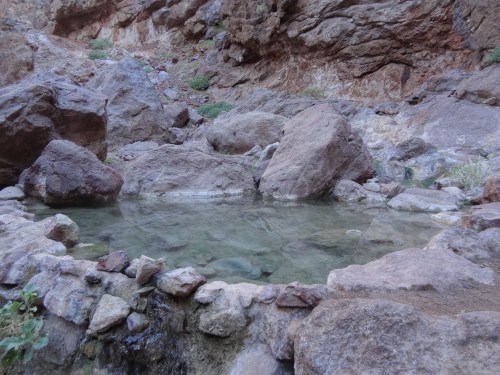 A natural hot spring surrounded by large rocks and a rocky landscape, with clear water reflecting the surroundings.