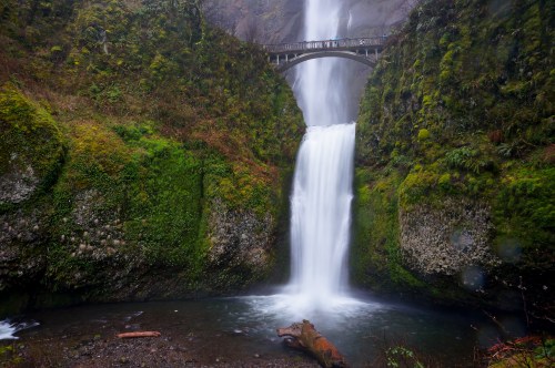 A majestic waterfall cascades down rocky cliffs, with a bridge above surrounded by lush green vegetation.