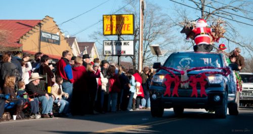 A festive parade with a decorated vehicle featuring Santa, surrounded by a crowd enjoying the event.