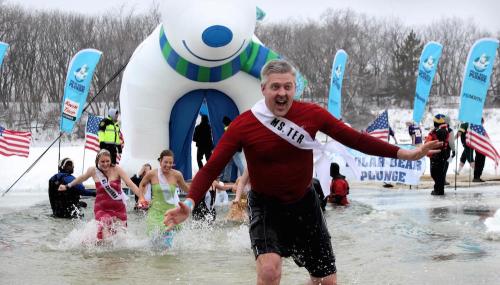 Participants in winter attire plunge into icy water, with a large polar bear inflatable and flags in the background.