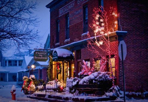 A charming brick building decorated with holiday lights and snow, featuring a flower shop and festive greenery.