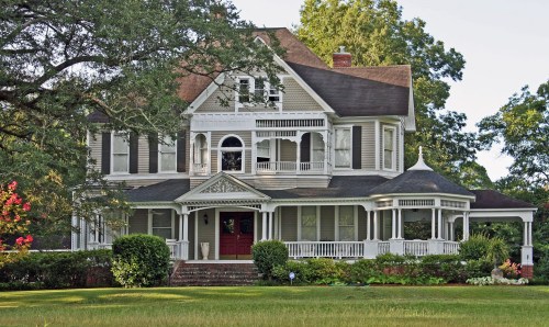 A large, elegant Victorian-style house with a wraparound porch and intricate architectural details, surrounded by greenery.