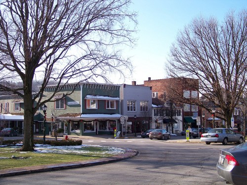 A quaint town square with shops, bare trees, and parked cars on a sunny winter day. Snow is visible on the ground.
