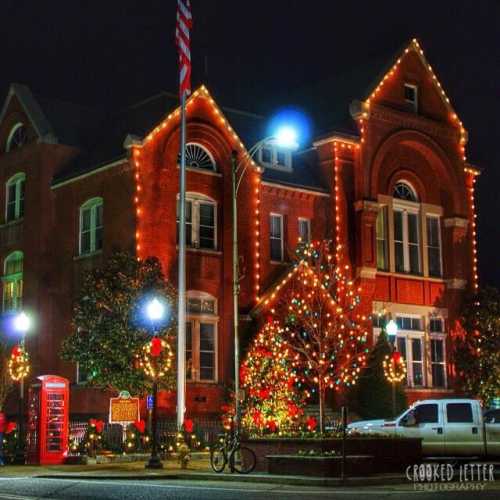 A festive building adorned with Christmas lights, surrounded by decorated trees and a vintage phone booth at night.