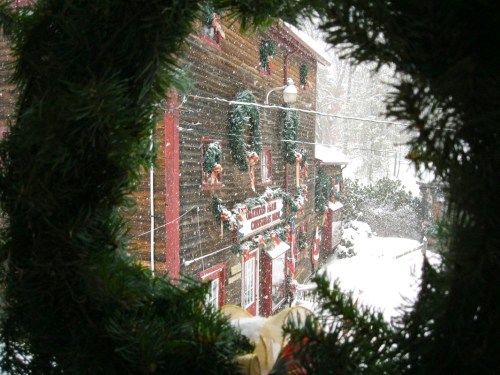 A snowy scene viewed through a festive wreath, showcasing a charming wooden building decorated for Christmas.