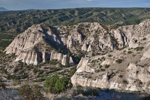 A scenic view of rugged, layered hills and valleys, covered in sparse vegetation under a clear blue sky.