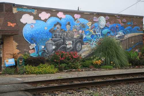 Colorful mural depicting two men in a vintage car, surrounded by whimsical clouds and flowers, on a brick wall.