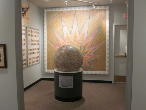A large, colorful mosaic wall with a spherical sculpture in the foreground, displayed in a gallery setting.
