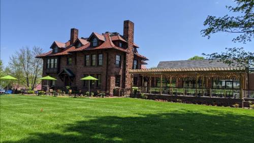 A large brick mansion with a red roof, surrounded by green grass and patio seating under umbrellas on a sunny day.