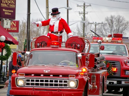 Santa Claus waves from a vintage fire truck during a festive parade, surrounded by holiday decorations.