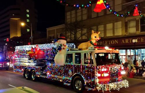 A festive fire truck decorated with lights, featuring a snowman, reindeer, and holiday decorations, driving in a parade.