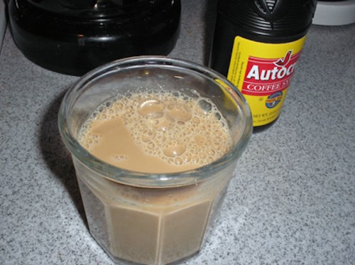 A glass of coffee with cream, bubbles on top, next to a bottle of coffee syrup on a countertop.