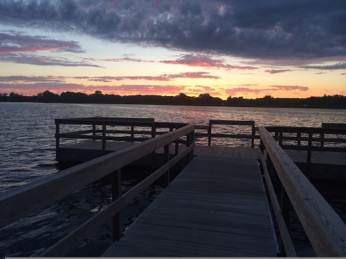 A wooden dock extends over a calm lake at sunset, with colorful clouds reflecting on the water.