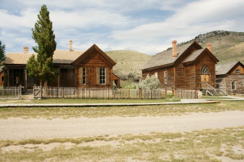 Historic wooden buildings in a ghost town, surrounded by grassy fields and mountains under a cloudy sky.