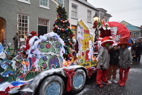 A festive parade float decorated with colorful lights and holiday themes, with children in winter attire posing nearby.