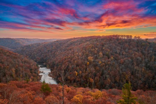 A vibrant sunset over a valley with colorful autumn foliage and a winding river below.