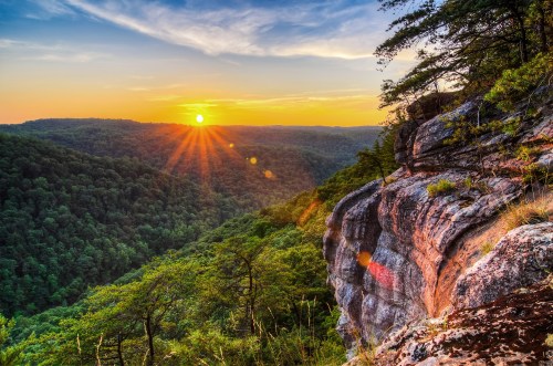 A vibrant sunset over a lush green valley, with rocky cliffs in the foreground and rays of sunlight shining through.
