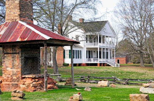 Historic white house with a porch, surrounded by trees and a stone chimney in the foreground. Green grass and a rustic fence.