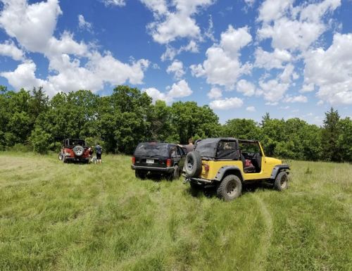 Two off-road vehicles parked in a grassy field under a blue sky with fluffy clouds, surrounded by trees.