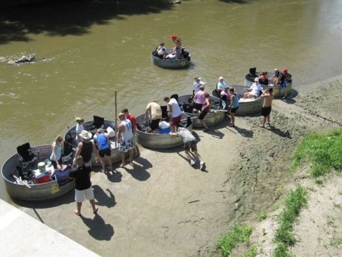 People gather around several small boats on a riverbank, enjoying a sunny day by the water.