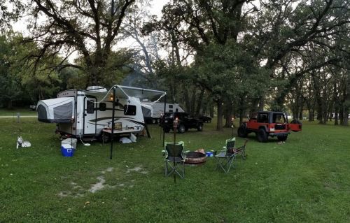 A campsite with a camper, two vehicles, and chairs set up around a fire pit under trees in a grassy area.