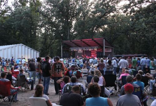 A large crowd gathers in a wooded area for an outdoor concert, with a stage illuminated by colorful lights.
