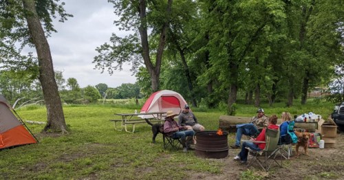 A group of people relaxes around a campfire at a campsite with tents and trees in the background.