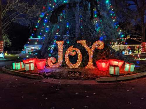 Colorful holiday lights illuminate the word "JOY" surrounded by glowing gift boxes in a festive outdoor display.