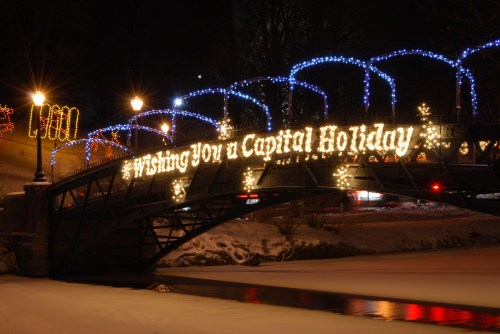 A bridge adorned with festive lights at night, displaying "Wishing You a Capital Holiday" over a snowy scene.