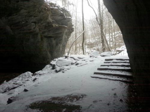 A snowy cave entrance with stone steps leading up, surrounded by rocky walls and bare trees in the background.