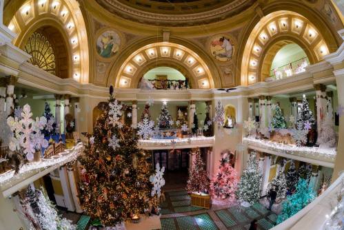 A festive hall decorated with numerous Christmas trees and snowflake ornaments, illuminated with warm lights.