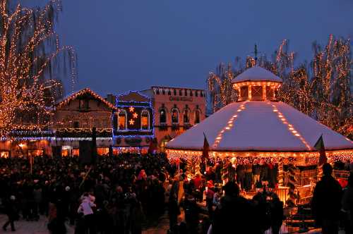 A festive scene with a crowd gathered around a lit gazebo, surrounded by buildings adorned with holiday lights at dusk.