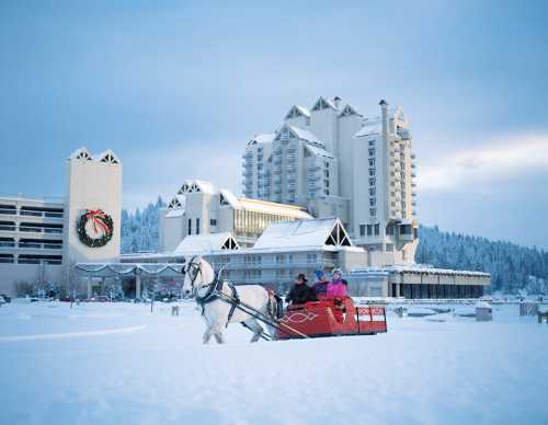 A horse-drawn sleigh rides through snow in front of a large hotel decorated for the holidays.