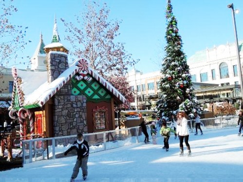 Children ice skate in a festive outdoor area with a decorated Christmas tree and a gingerbread house.