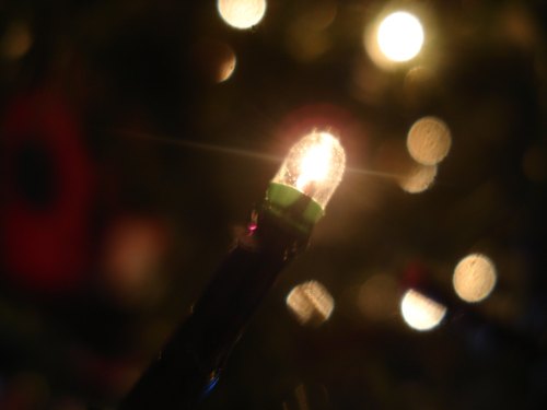 A close-up of a glowing Christmas light bulb with blurred festive lights in the background.