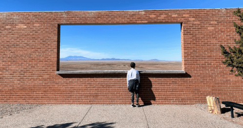 A child stands by a brick wall with a large window, gazing at a vast landscape and distant mountains.