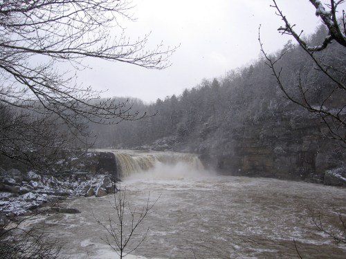A snowy landscape featuring a waterfall cascading into a river, surrounded by trees and rocky cliffs.