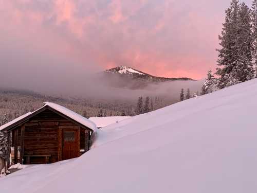 A rustic cabin surrounded by deep snow, with a mountain peak and pink-hued clouds in the background at sunset.