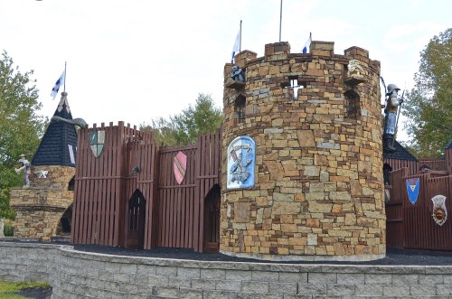A stone castle with wooden walls, flags, and shields, surrounded by trees under a cloudy sky.