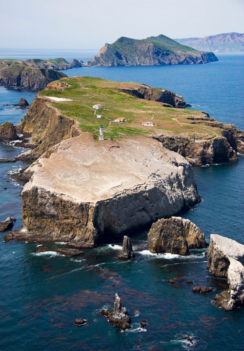 Aerial view of a rugged island with a lighthouse, surrounded by blue ocean and distant mountains.