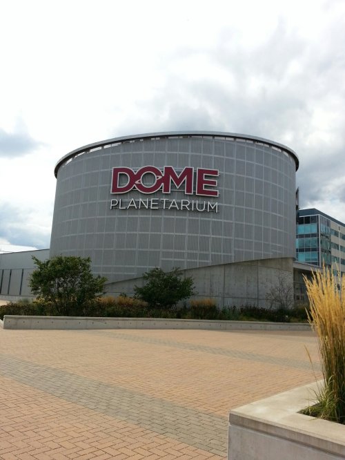 Exterior of a modern planetarium building with a large sign reading "DOME PLANETARIUM" against a cloudy sky.