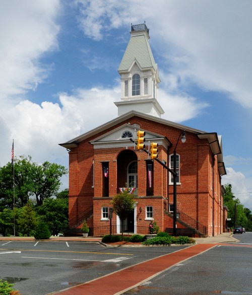 Historic brick building with a tall clock tower, surrounded by trees and a blue sky with clouds.