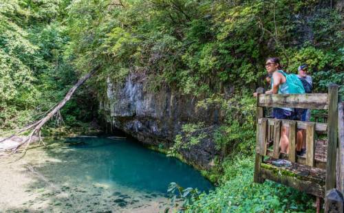 A person stands on a wooden platform by a serene, green-tinted pond surrounded by lush trees and rocky cliffs.