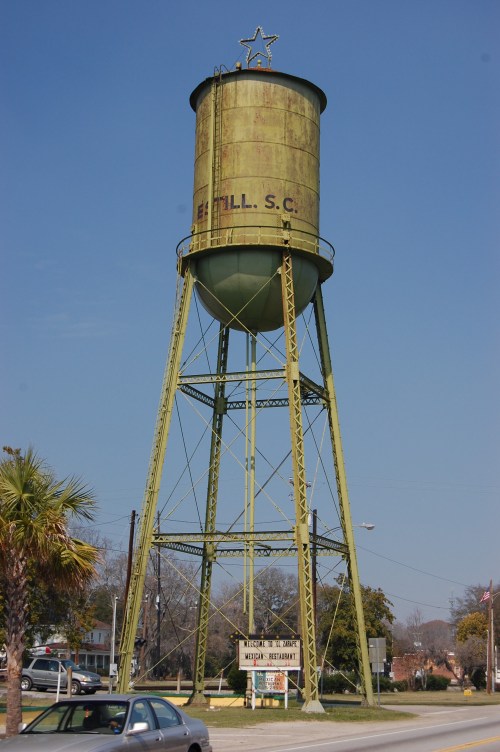 A tall, green water tower with a star on top, standing beside a road and a restaurant sign in a small town.