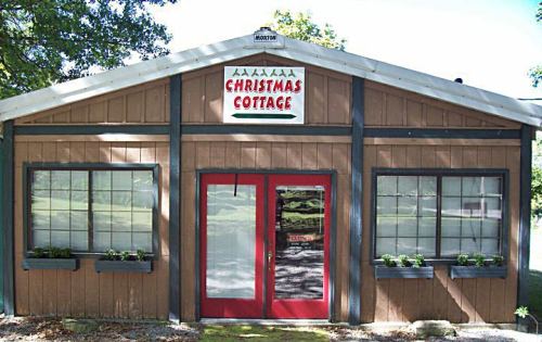 A cozy wooden building with a sign reading "Christmas Cottage" and red doors, surrounded by greenery.